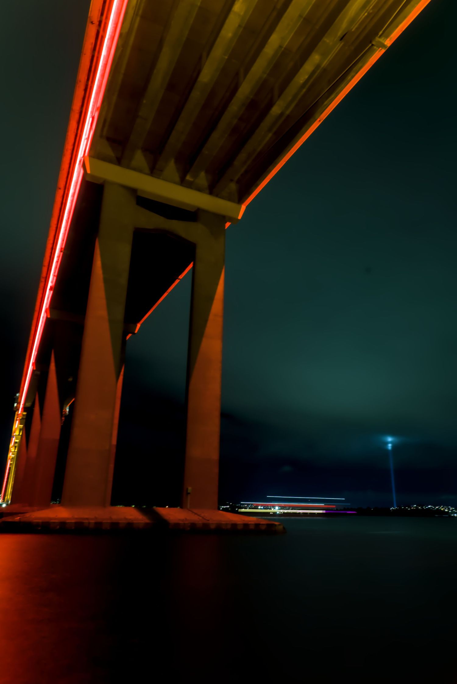 Night view under an illuminated orange bridge over water with red light trails and a distant glowing tower.