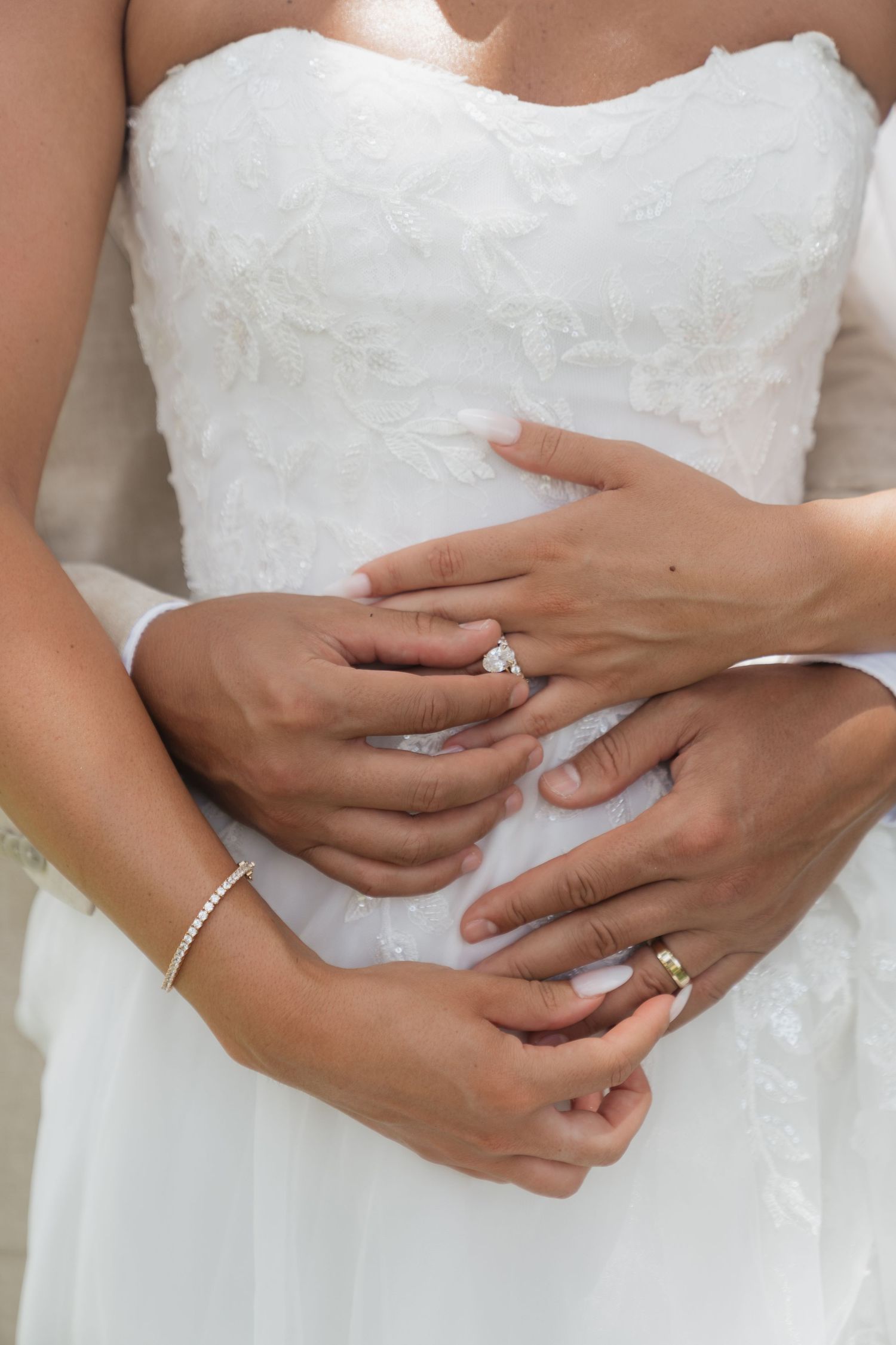Bride and groom hands intertwined showing engagement ring, wedding band, and diamond bracelet on white lace wedding dress.