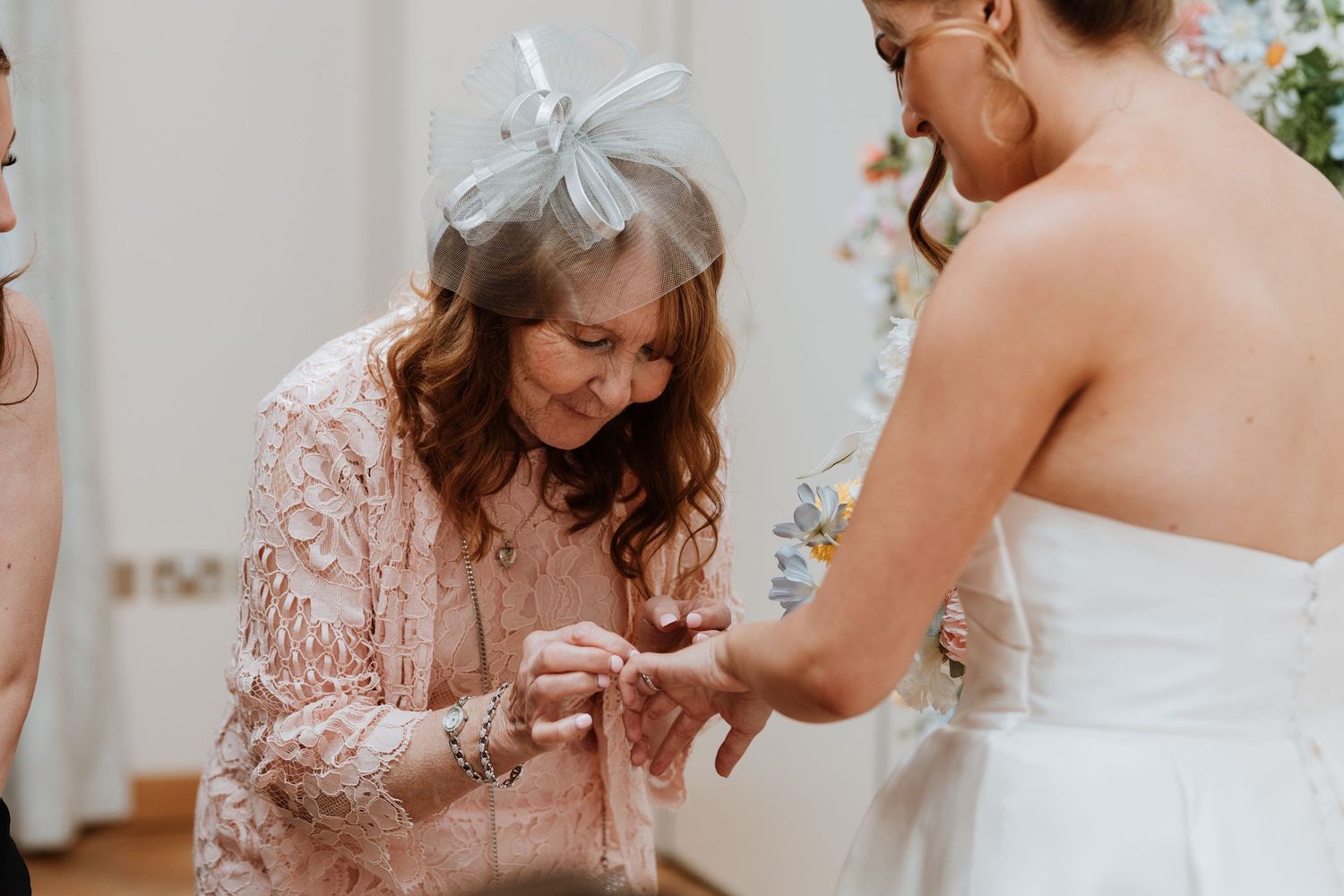 Older woman in pink lace dress and blue fascinator placing a ring on a bride's finger during wedding ceremony.