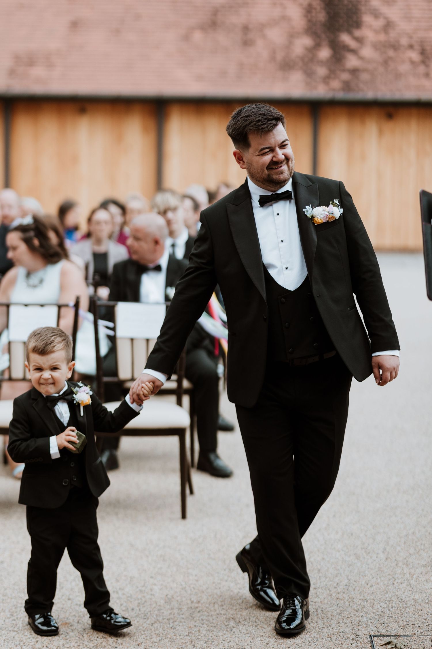 Groom in black tuxedo walks down wedding aisle holding hands with young ring bearer in matching suit.