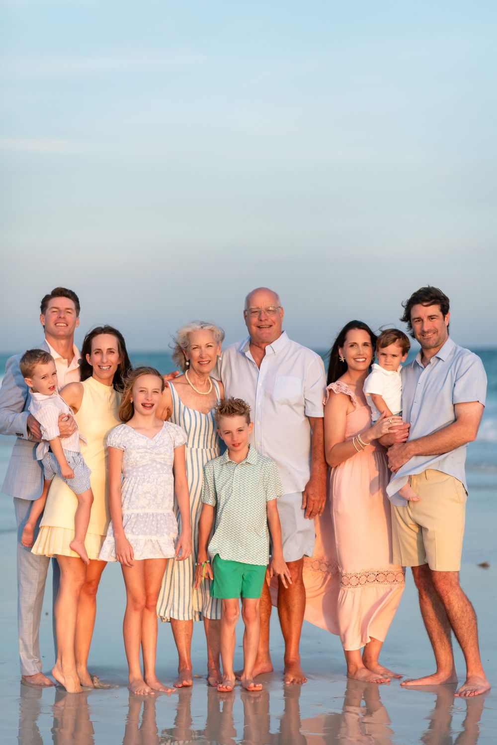 Multi-generational family of nine poses together on a beach at sunset, smiling in coordinated pastel outfits.