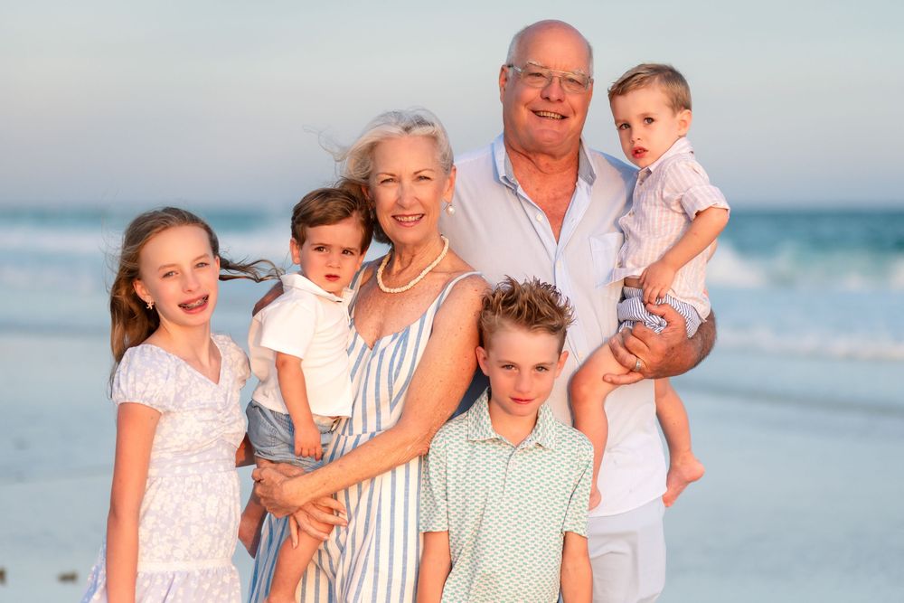 Grandparents smiling with four young grandchildren on a sandy beach at sunset in a warm family portrait.