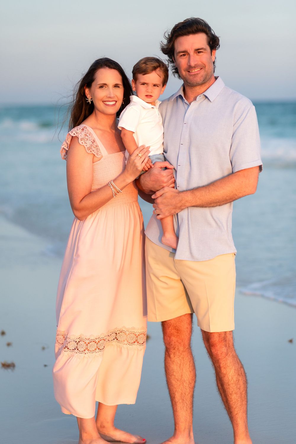 Happy family of three posing on a sandy beach at sunset, parents holding young child with ocean waves behind them.