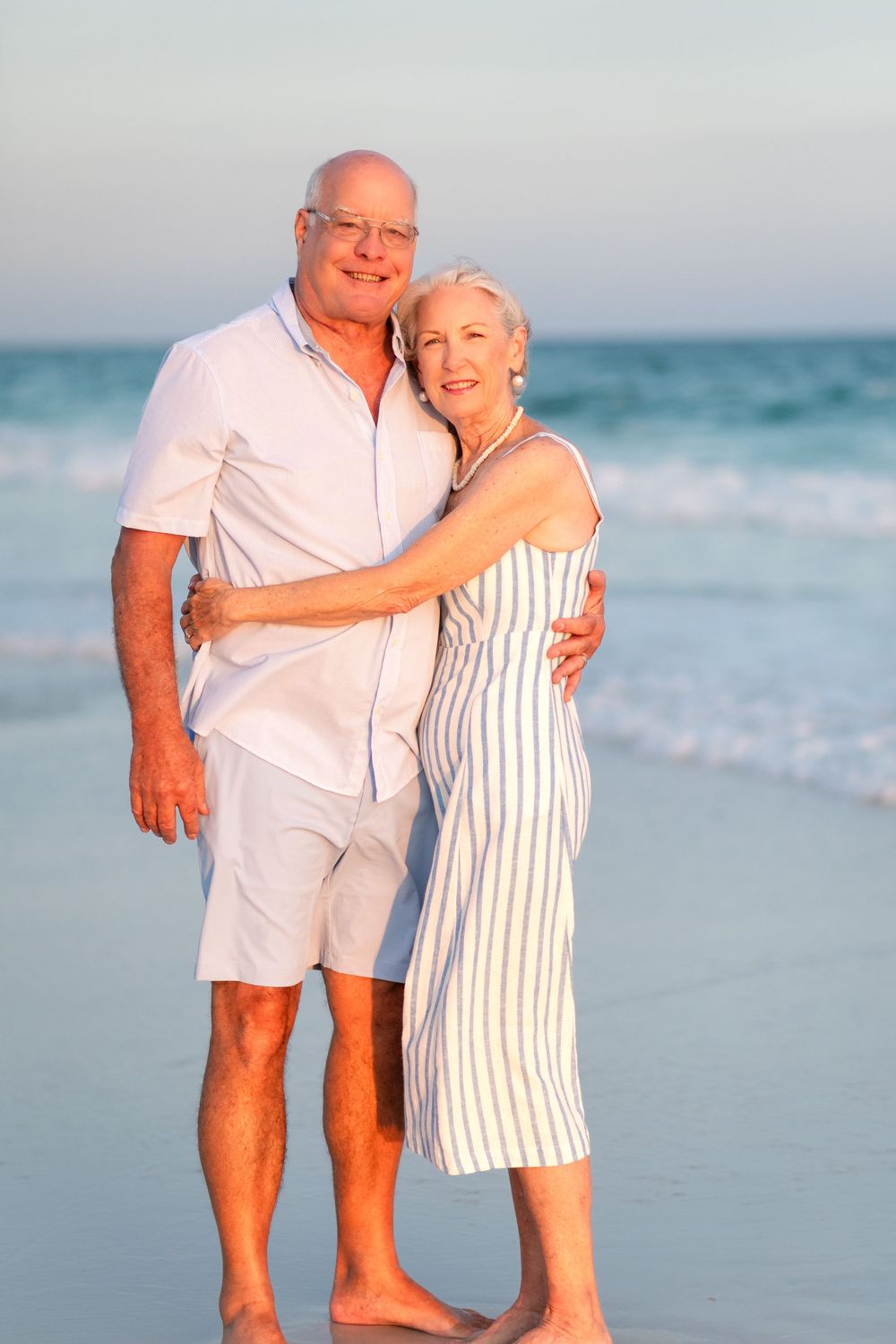 Senior couple embracing and smiling on a sunny beach, man in white shirt and shorts, woman in striped dress.
