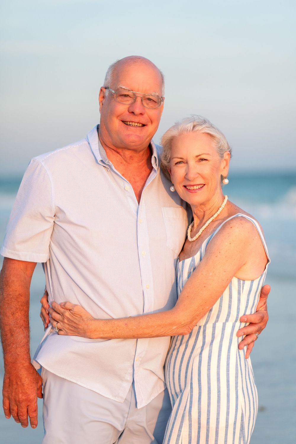 Happy senior couple embracing on a beach at sunset, wearing white and blue outfits, smiling warmly.