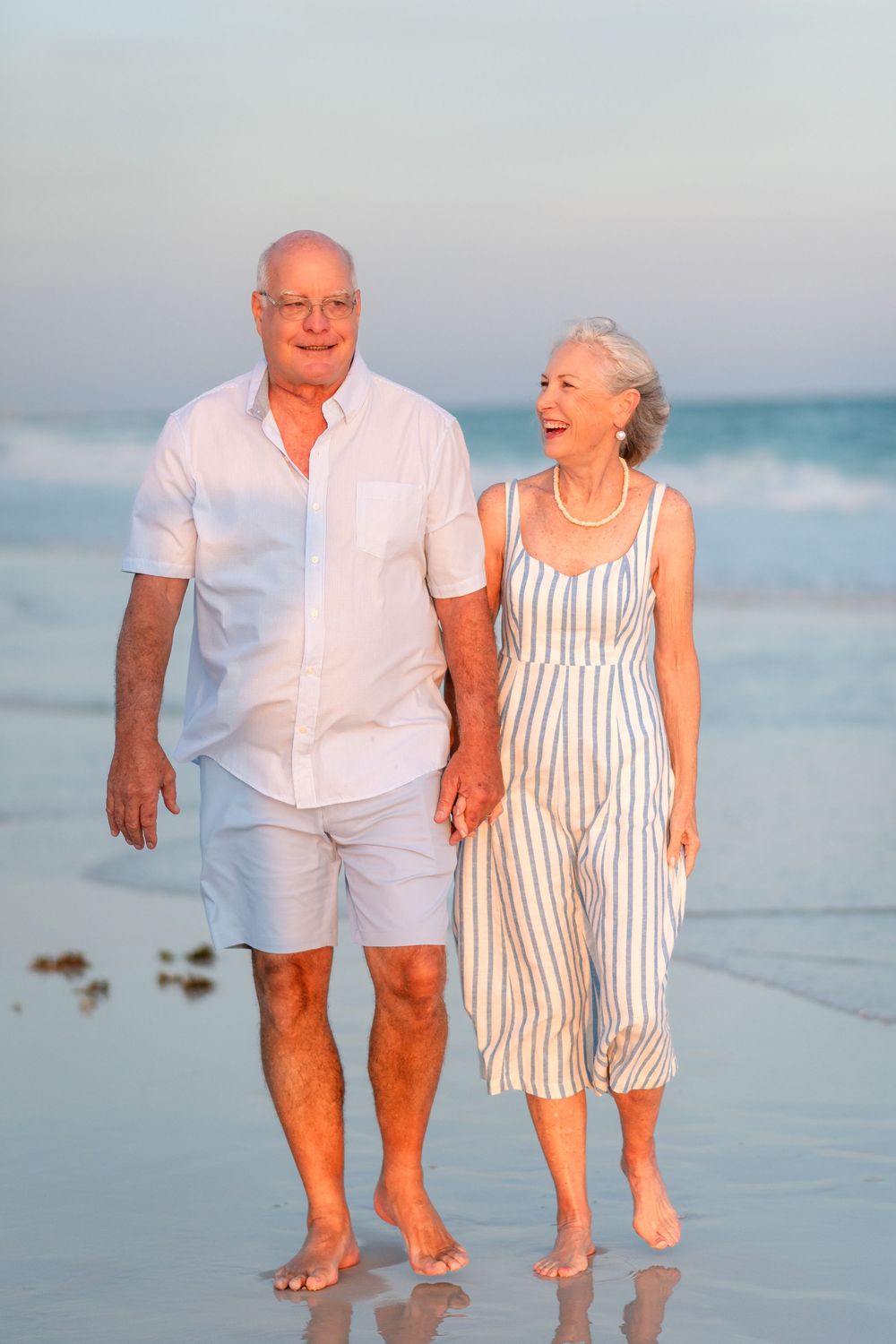 Senior couple walking barefoot on a sandy beach at sunset, smiling and enjoying a romantic stroll by the ocean.