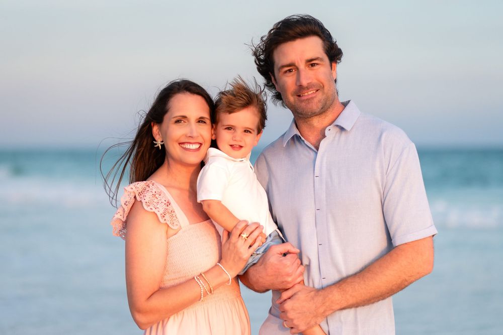 Happy family of three posing on a sunny beach, parents holding young toddler with ocean waves in background.