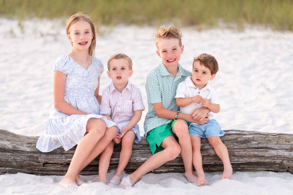 Four children sitting on a driftwood log on a white sand beach, smiling and posing for a family photo.