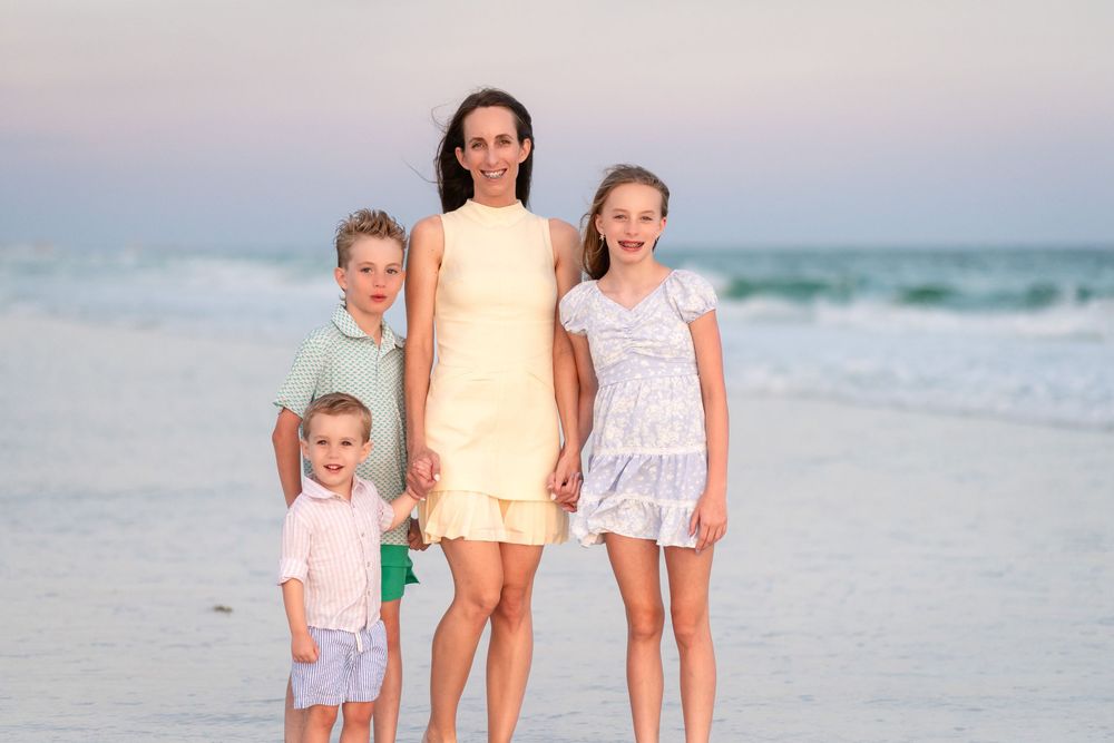Family of four posing on a beach at sunset, mother with three children smiling by the ocean shore.