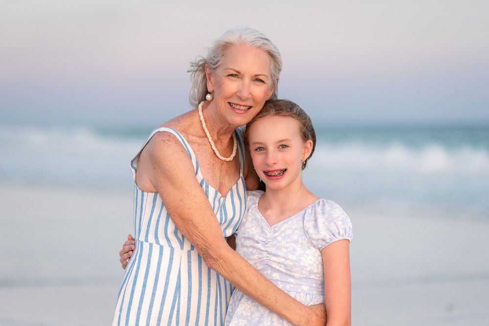 Grandmother and granddaughter smiling together on a beach at sunset, wearing summer dresses.