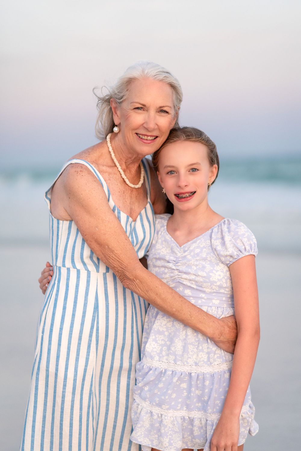 Grandmother in striped dress hugging granddaughter on beach at sunset, both smiling at camera.