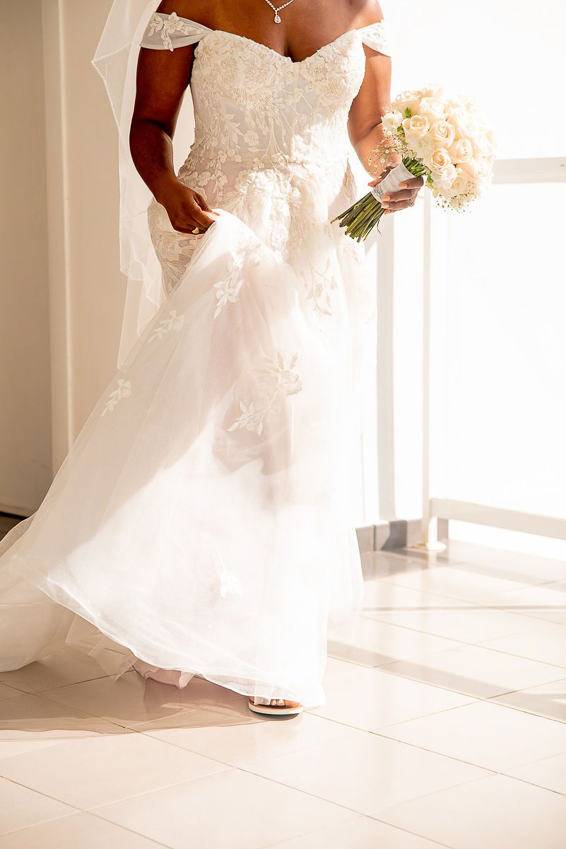 Bride in off-shoulder lace wedding gown holding white floral bouquet, standing near bright window in elegant bridal look.