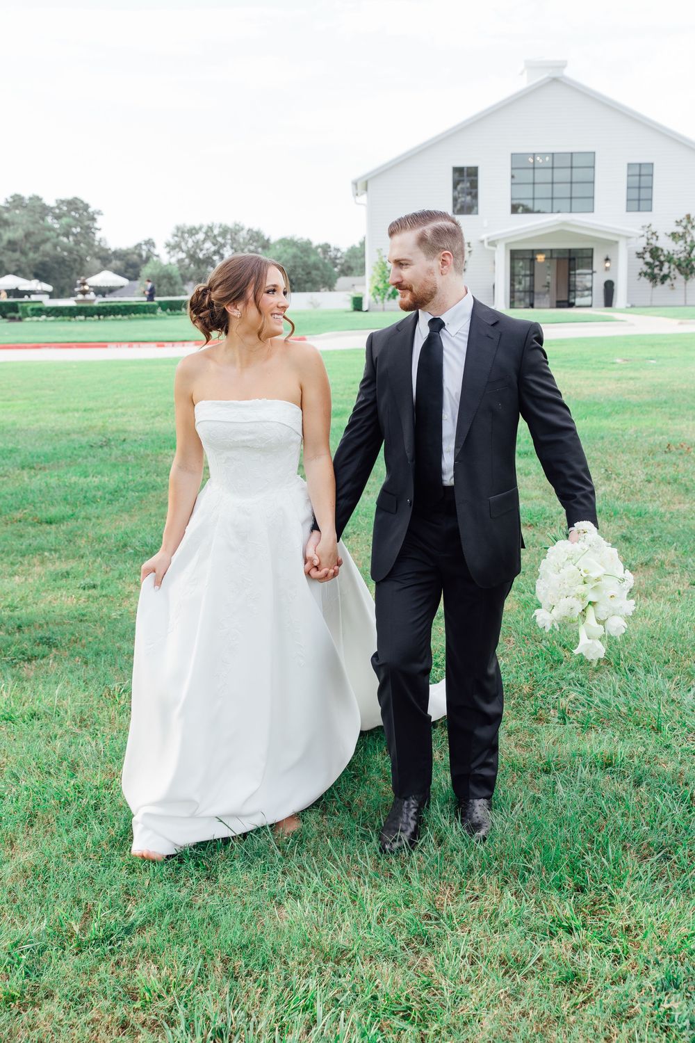 Bride and groom walking hand in hand on green lawn with white venue building in background.