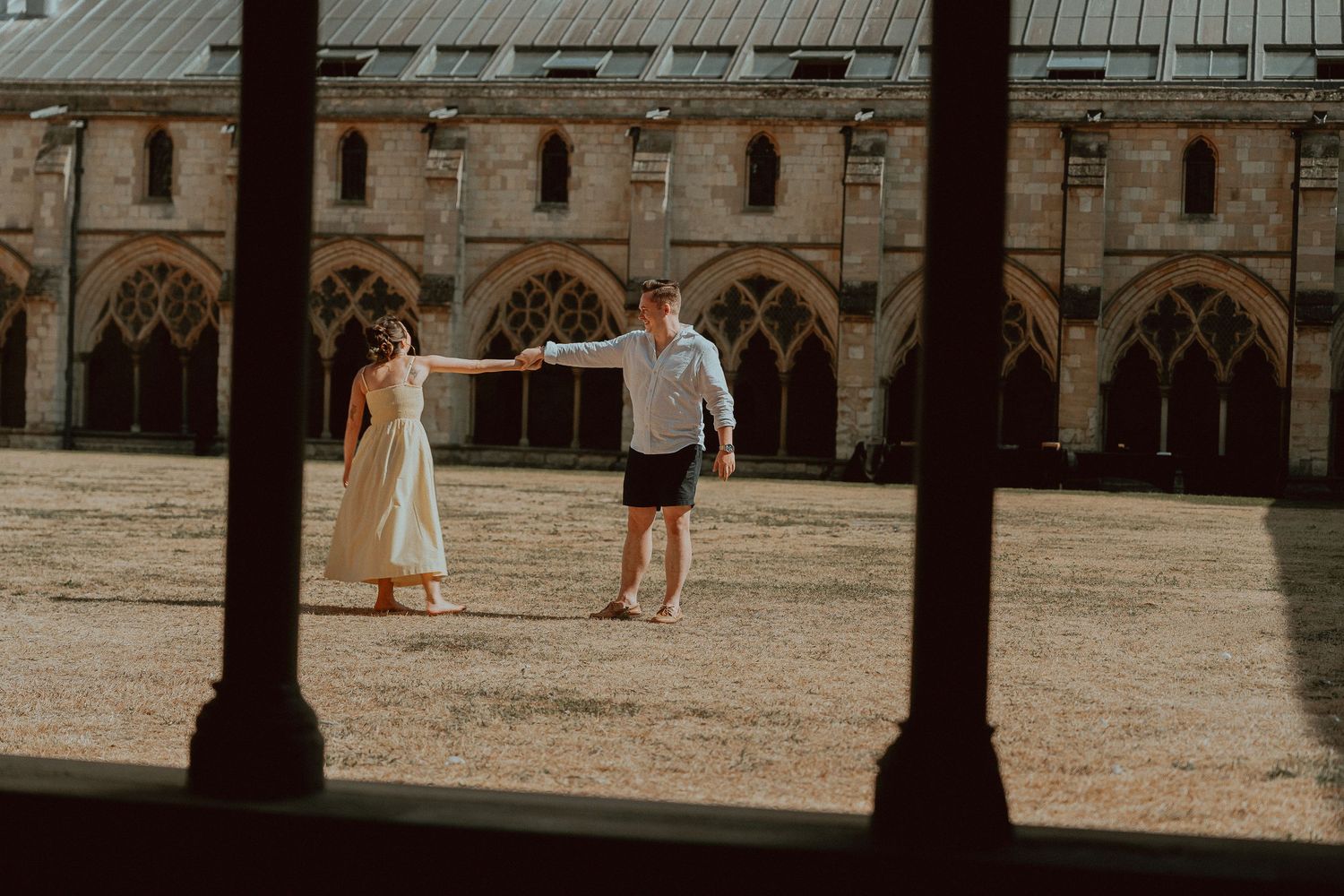 Couple holding hands in a sunlit historic stone courtyard with Gothic arched cloisters in the background.