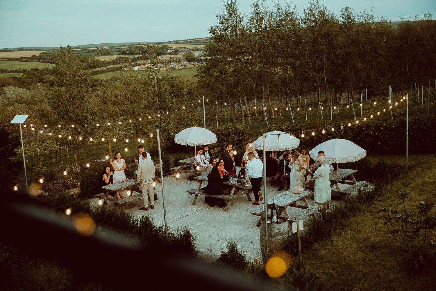 Outdoor evening gathering with string lights, white umbrellas, and guests socializing on a patio surrounded by lush greenery.