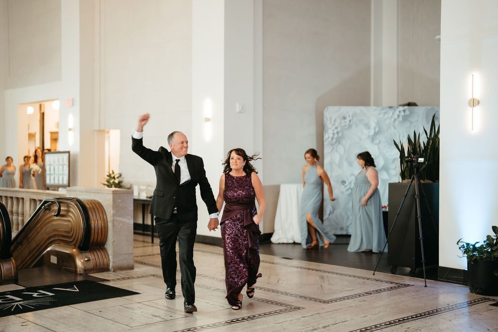 Couple makes grand entrance at elegant wedding reception venue, man raising fist in celebration, bridesmaids visible in background.