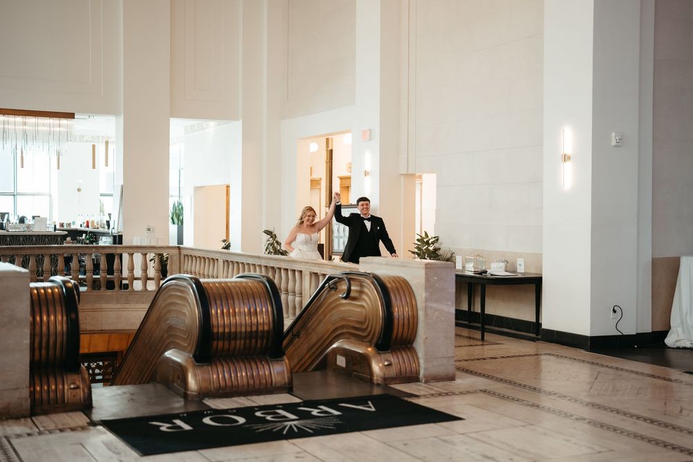 Bride and groom waving on escalator in elegant hotel lobby with marble floors and Art Deco decor.