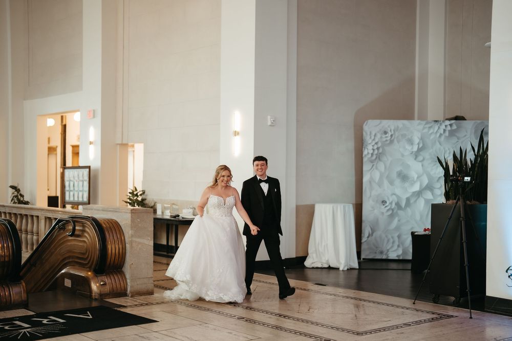 Bride and groom walk hand-in-hand through elegant wedding venue lobby with white floral backdrop.