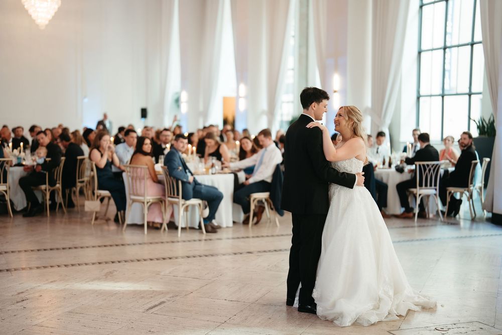 Bride and groom share their first dance in an elegant white ballroom surrounded by seated wedding guests.