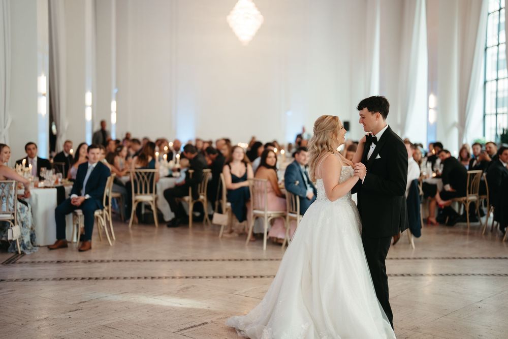Bride and groom share their first dance at elegant wedding reception with guests seated at gold chairs in bright ballroom.