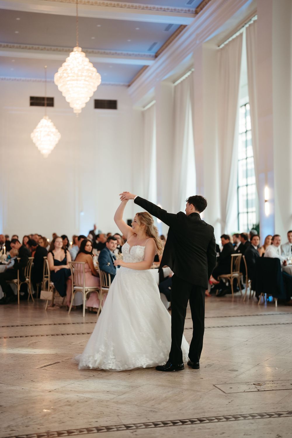 Bride and groom share their first dance in an elegant ballroom with chandeliers and guests seated around the dance floor.