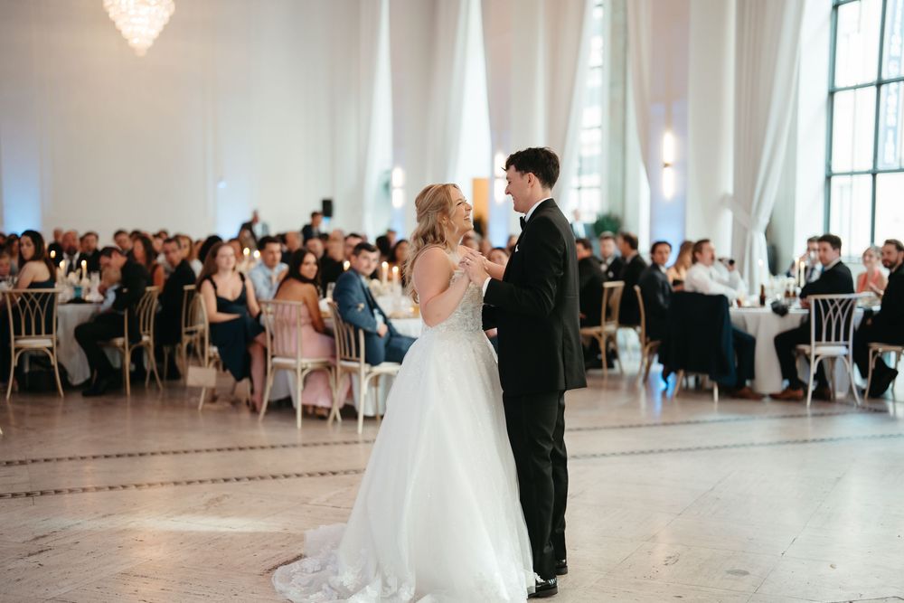 Bride and groom share their first dance in an elegant ballroom surrounded by seated wedding guests.