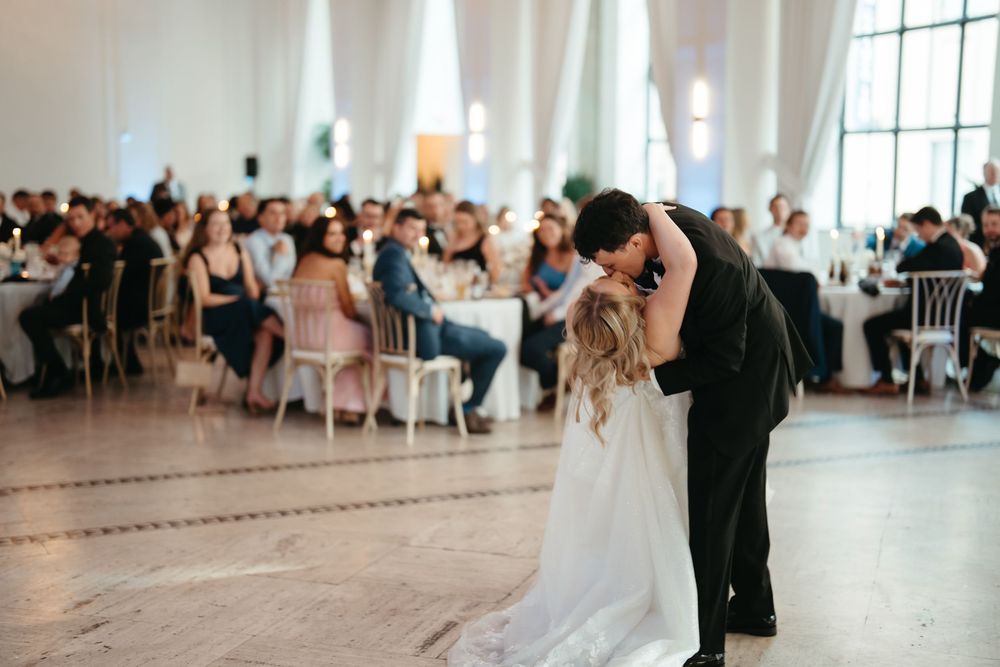 Bride and groom share first dance kiss at elegant wedding reception with seated guests in bright ballroom.