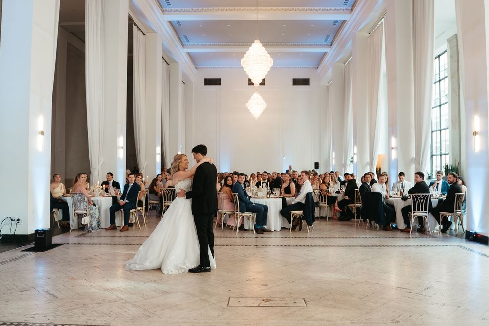 Bride and groom share first dance in elegant white ballroom with chandelier, surrounded by seated wedding guests.