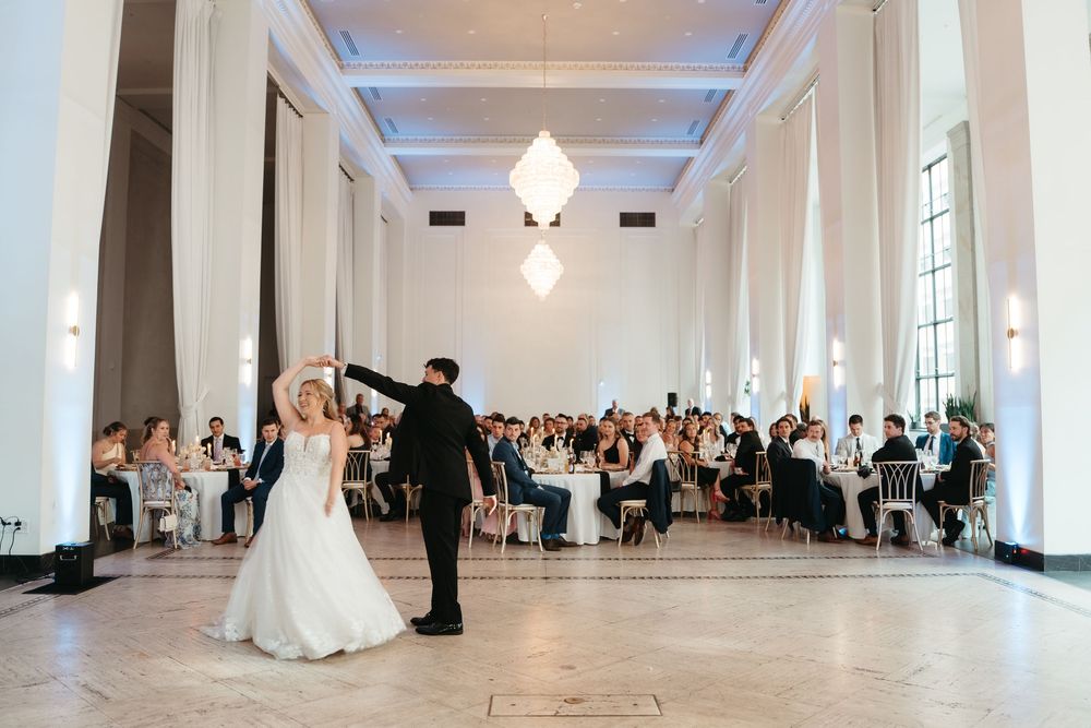 Bride and groom share first dance in elegant white ballroom with guests seated at reception tables behind them.