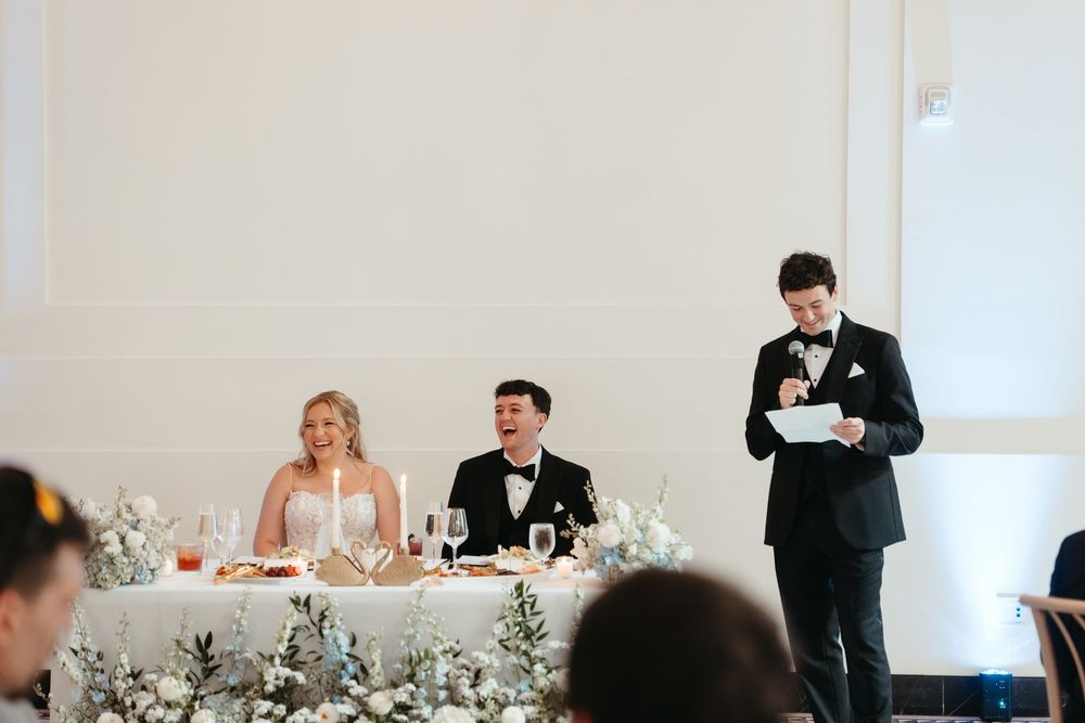 Bride and groom seated at wedding reception head table while best man delivers speech in black tuxedo.