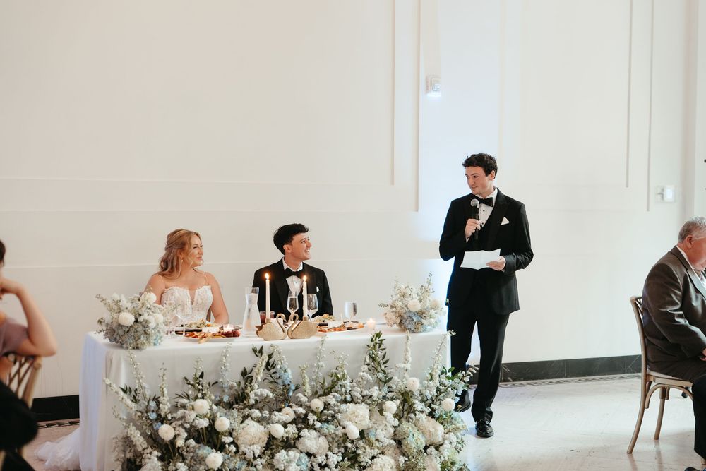 Groom giving wedding speech at elegant reception with floral arrangements and bride seated at sweetheart table.