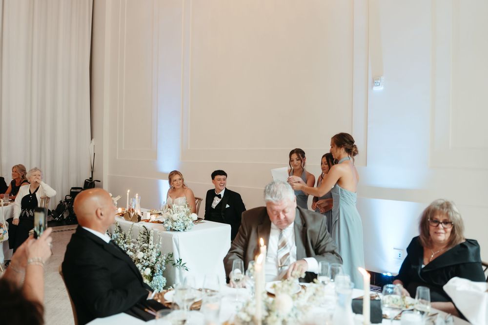 Bridesmaid giving a toast speech at an elegant wedding reception with guests seated at decorated tables.