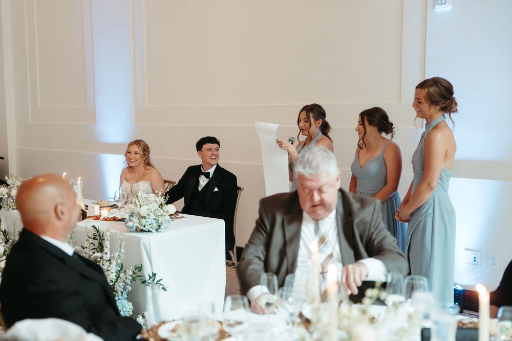Wedding reception scene with bride and groom at sweetheart table while bridesmaids in blue give toasts.