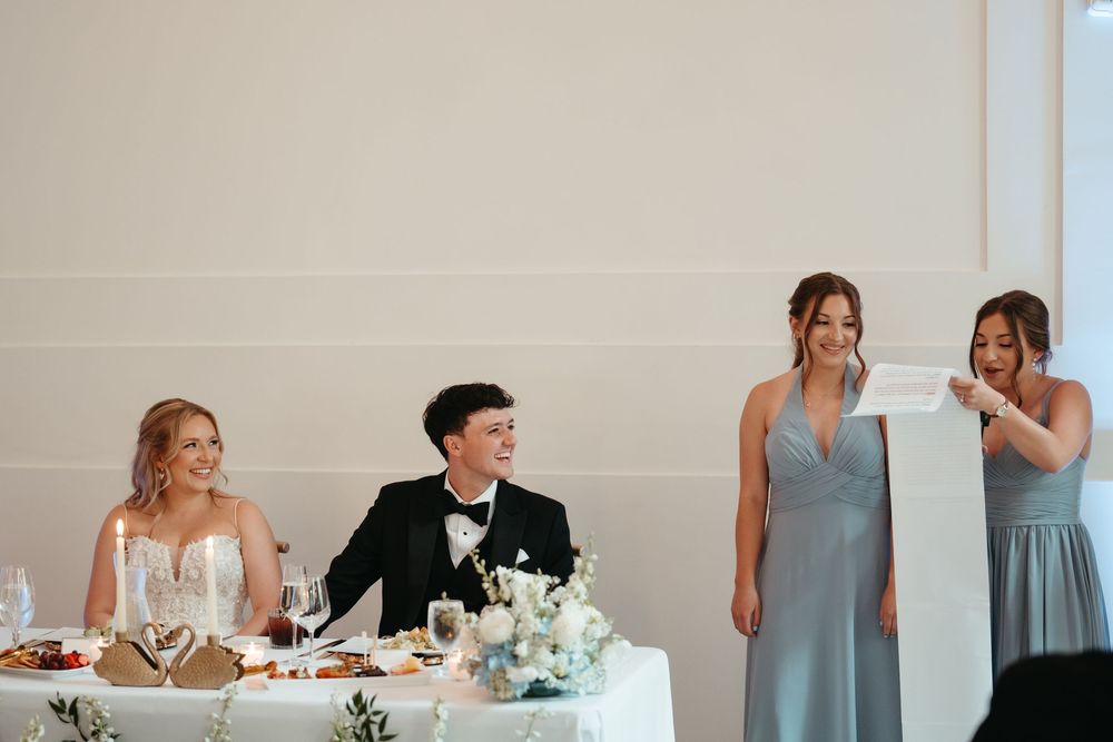 Bridesmaid gives a heartfelt speech at wedding reception while bride and groom smile at the head table.