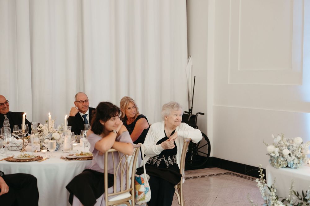 Elegant wedding reception guests seated at a decorated table with white floral arrangements and candles in a bright venue.