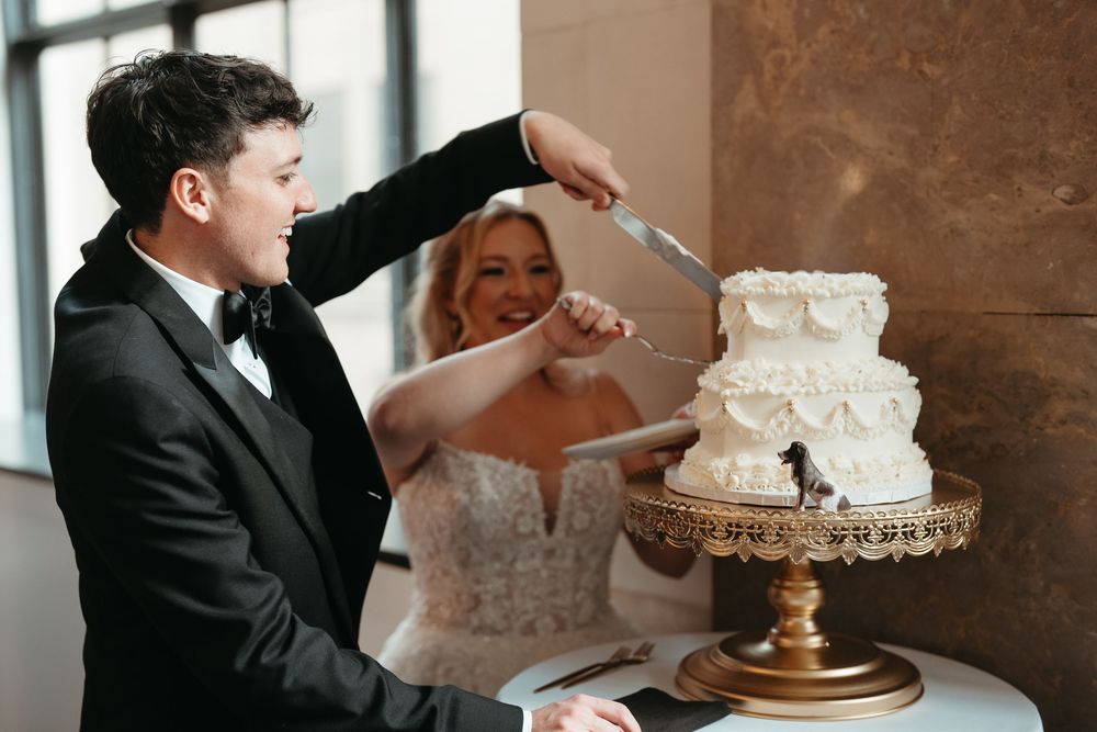 Smiling groom cuts elegant white tiered wedding cake with bride at elegant wedding reception.
