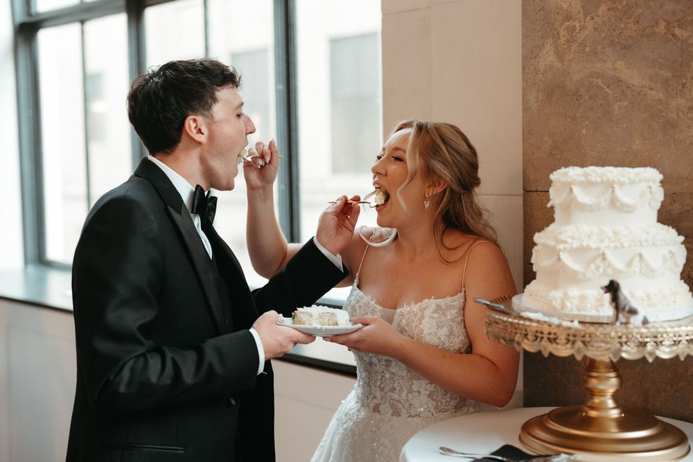 Bride and groom feeding each other wedding cake at their elegant reception, with a white tiered cake on a gold stand.