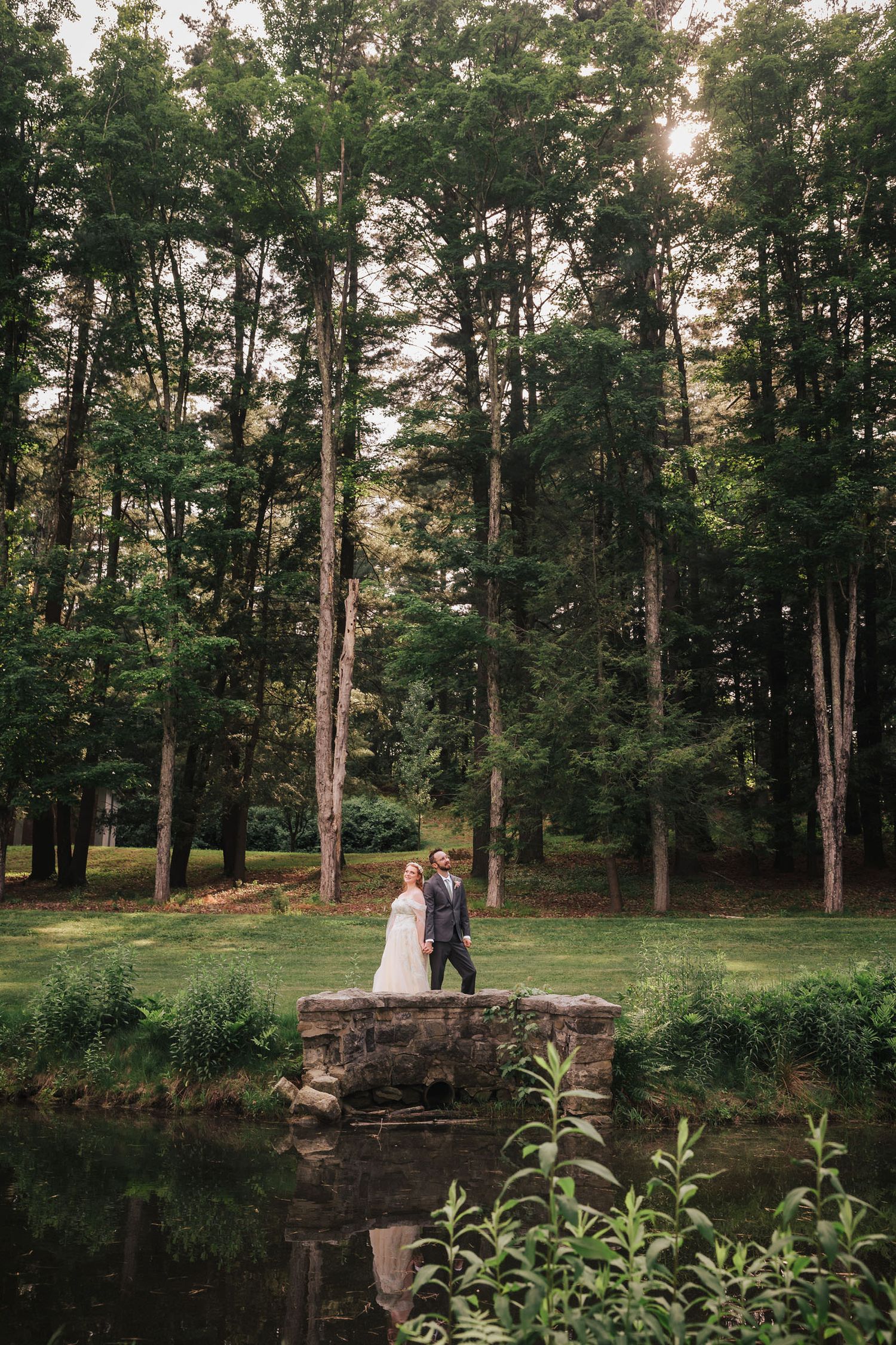 Bride and groom standing on a stone footbridge over a pond at golden hour, tall pines reflected in the still water below at Searles Castle.