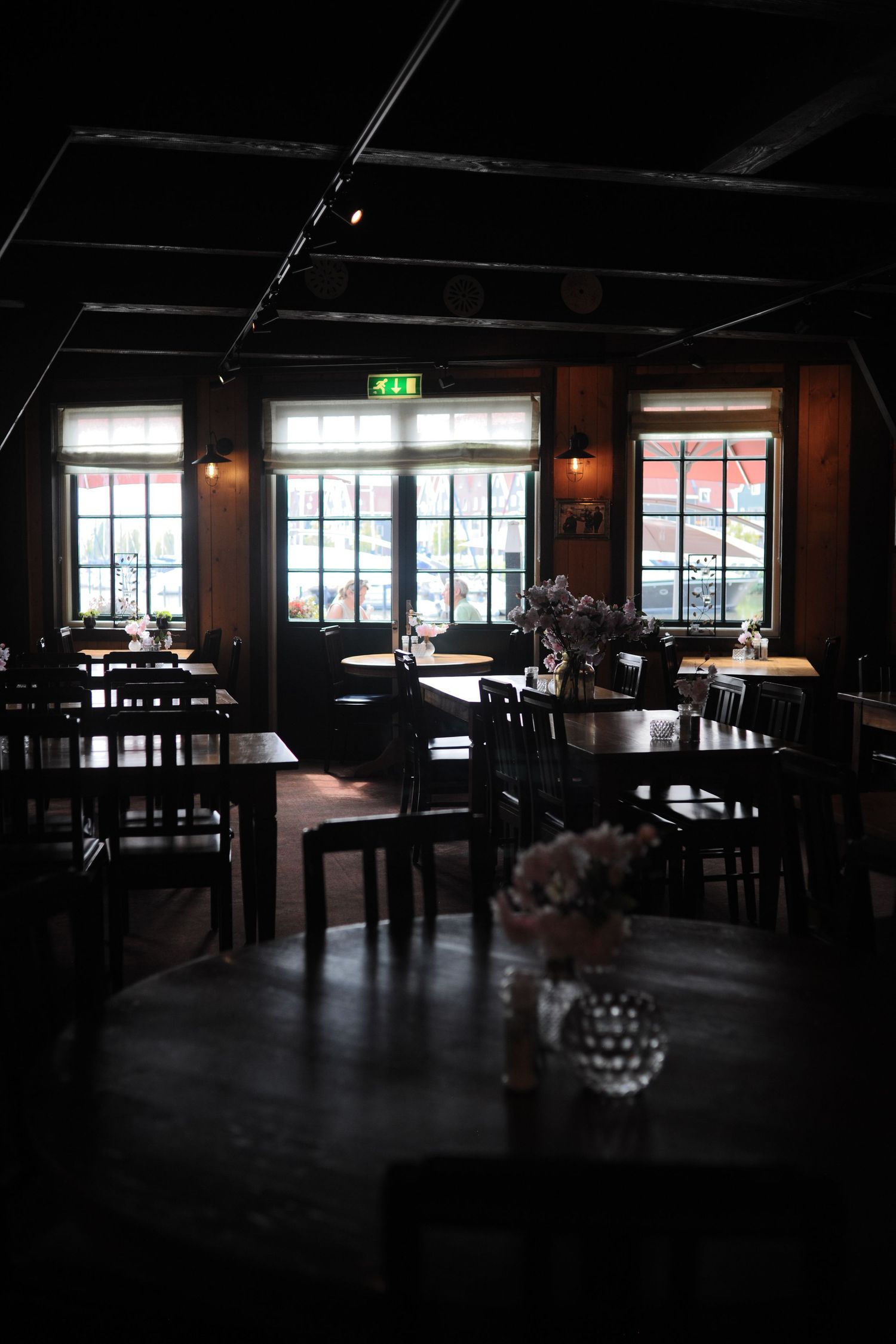Dimly lit restaurant interior with dark wood tables, chairs, and large windows with an exit sign glowing above.