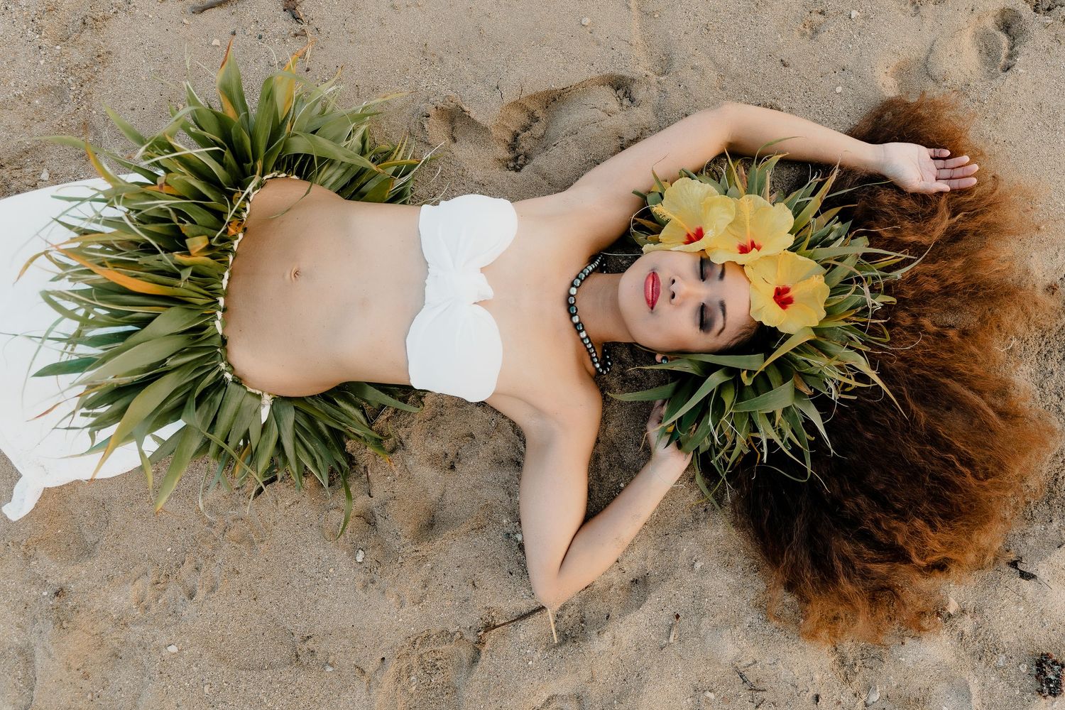 Young woman in floral dress and tropical flower crown poses by ocean rocks with long curly hair flowing.