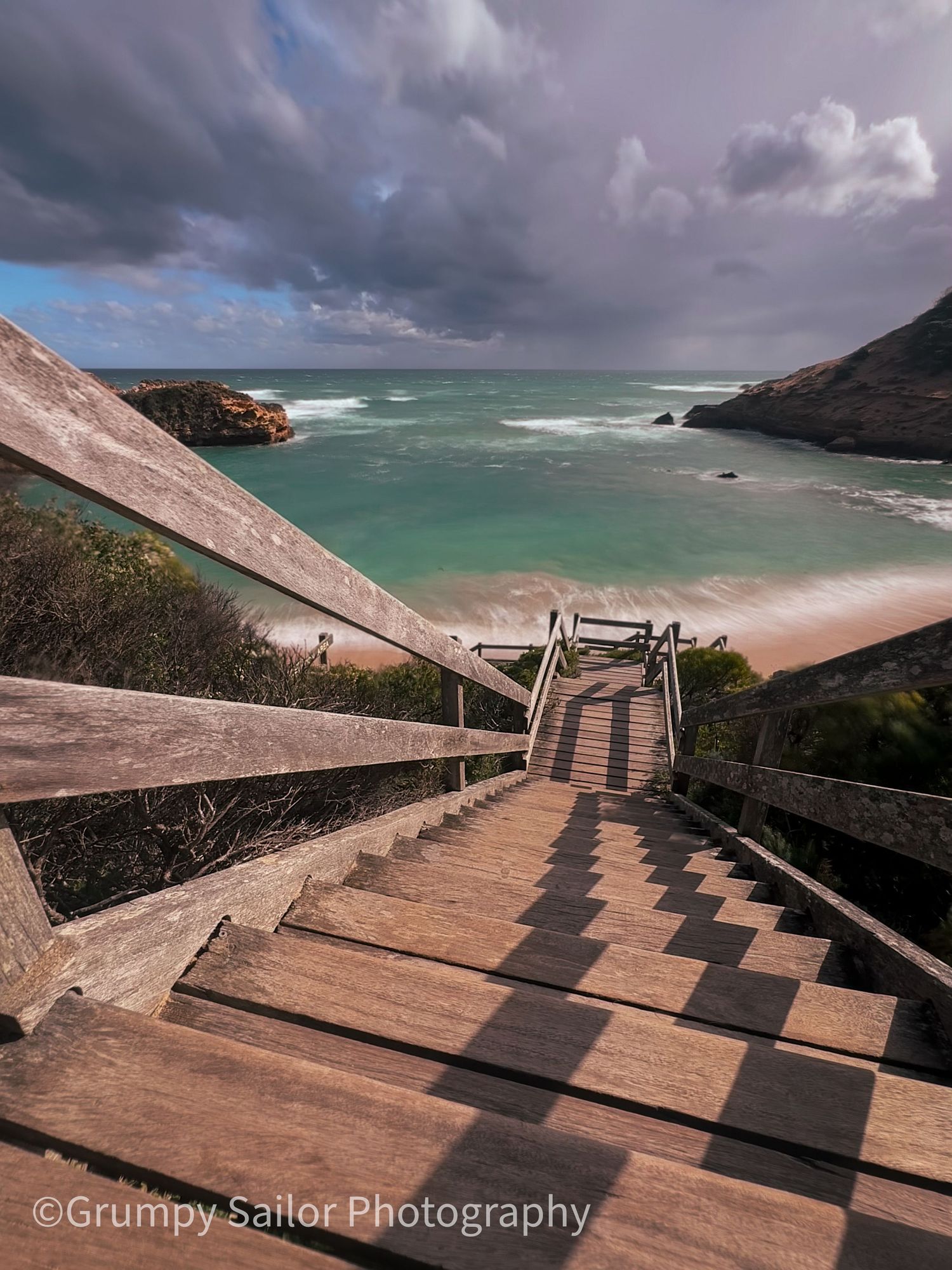 Wooden staircase descending to a turquoise beach under dramatic storm clouds, coastal landscape photography.