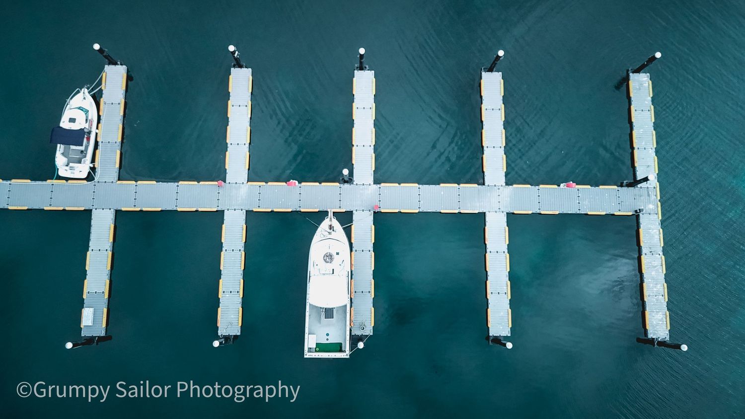 Aerial view of floating marina docks forming a comb pattern with two white boats moored in turquoise water.