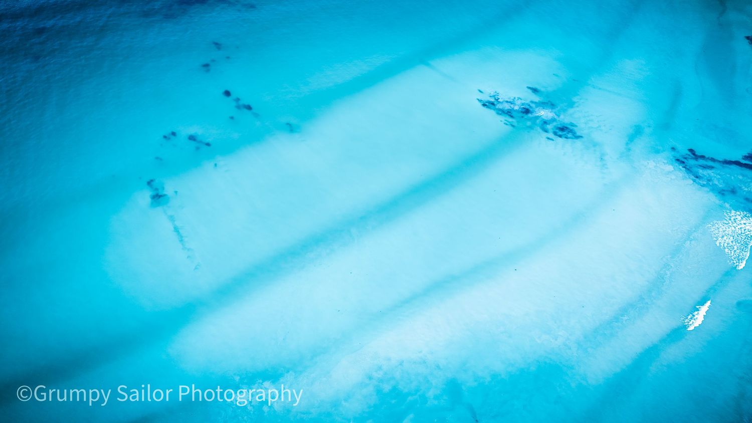 Aerial view of shallow turquoise ocean water revealing sandy seabed patterns and gentle wave shadows.