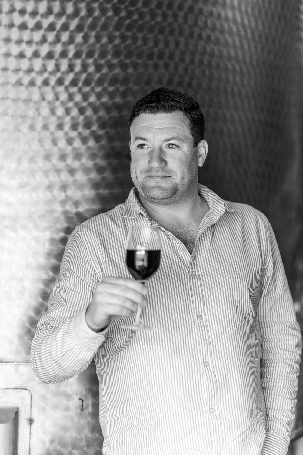 Black and white photo of a man in a striped shirt holding a glass of red wine in a winery cellar.