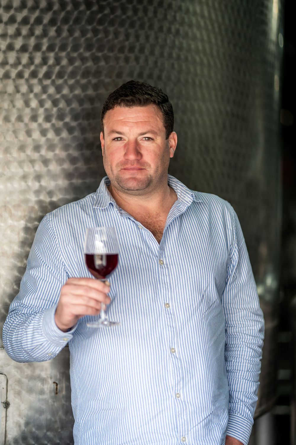 Man holding a glass of red wine standing in front of stainless steel fermentation tanks in a winery.