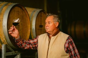 A winemaker examines a glass of red wine in a barrel cellar, wearing a plaid shirt and tan vest.