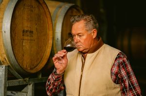 A winemaker carefully smells a glass of red wine in a barrel cellar, wearing a plaid shirt and vest.