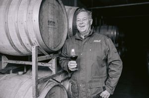 Man smiling holding red wine glass beside oak barrels in winery cellar, black and white photo.