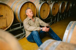 Man relaxing against wine barrels in a cellar, holding a glass of red wine, wearing a beige shirt and jeans.