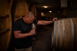Man smelling wine glass while leaning on oak barrel in rustic wine cellar during tasting.